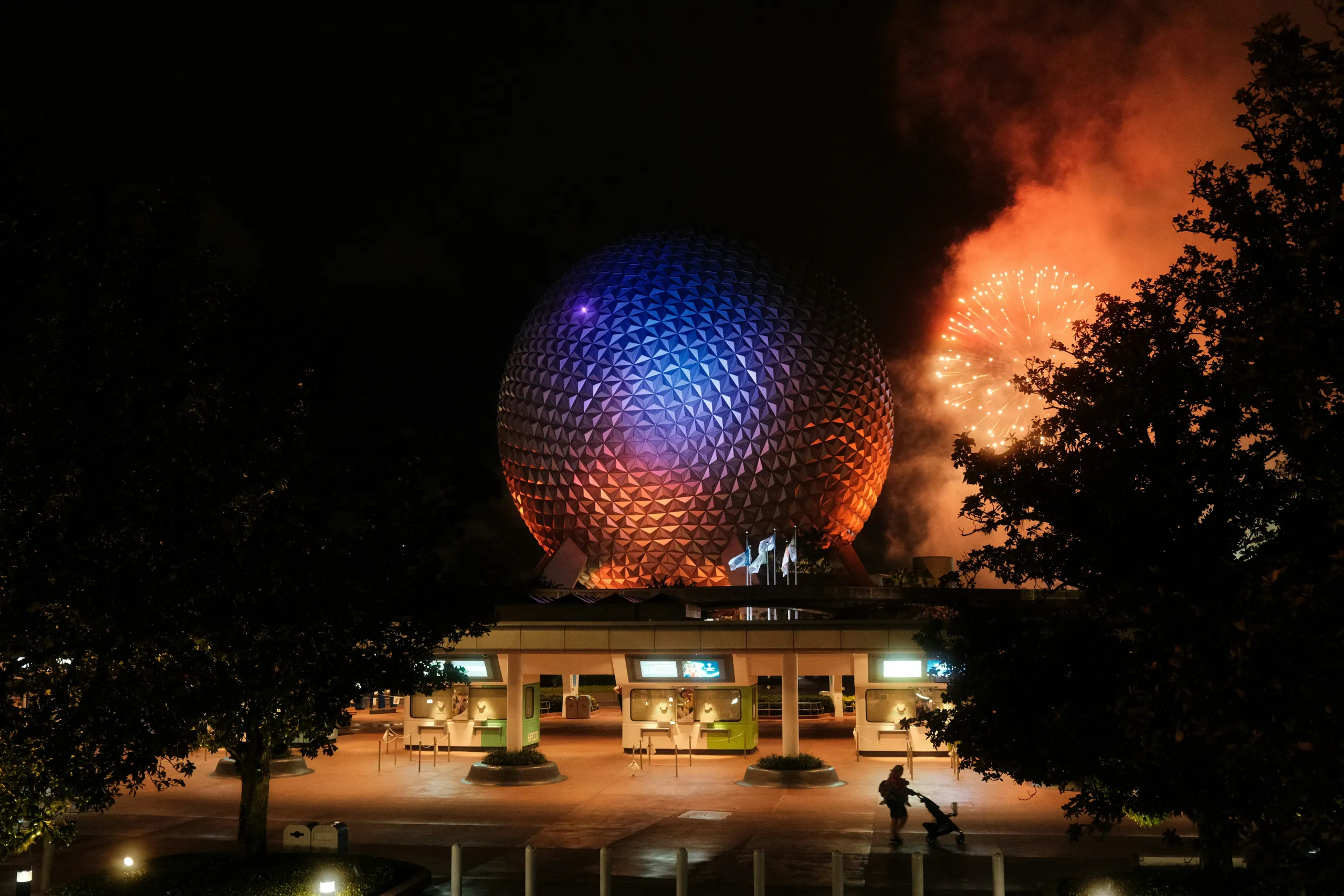 Spaceship Earth at EPCOT glowing blue and orange at night, with fireworks behind the trees